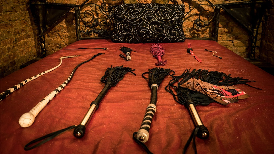 Bed with red bedspread and several whips, floggers, and impact toys arranged on it