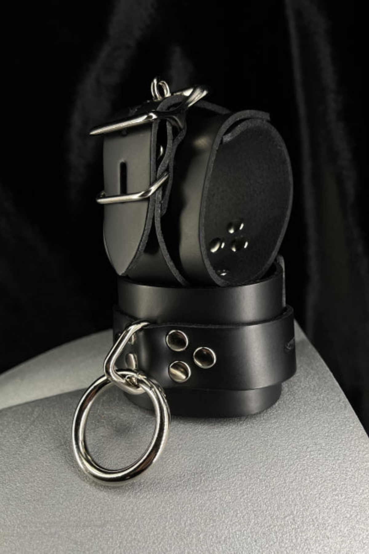 Black leather wrist cuffs with a locking buckle and silver O-ring, stacked on a grey mannequin form in front of draped black velvet.