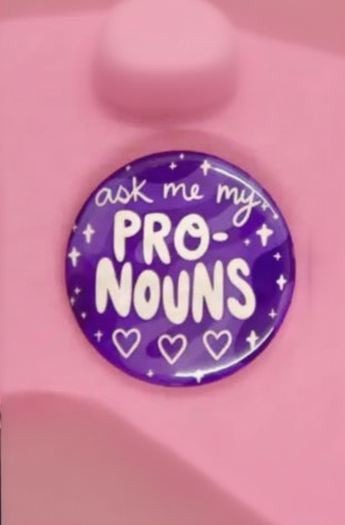 Purple Ask Me My Pronouns pinback button with white PRO-NOUNS lettering, sparkles and three hearts on a soft pink background