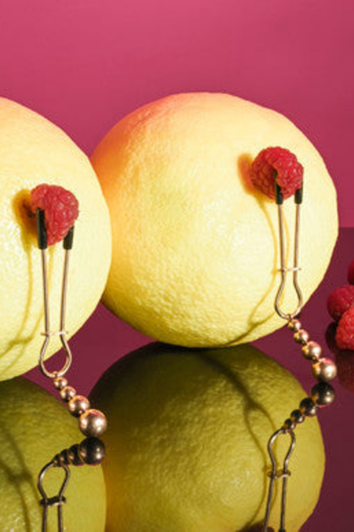Two rose gold tweezer nipple clamps gripping raspberries on yellow fruit against pink, showing the beaded weights hanging below