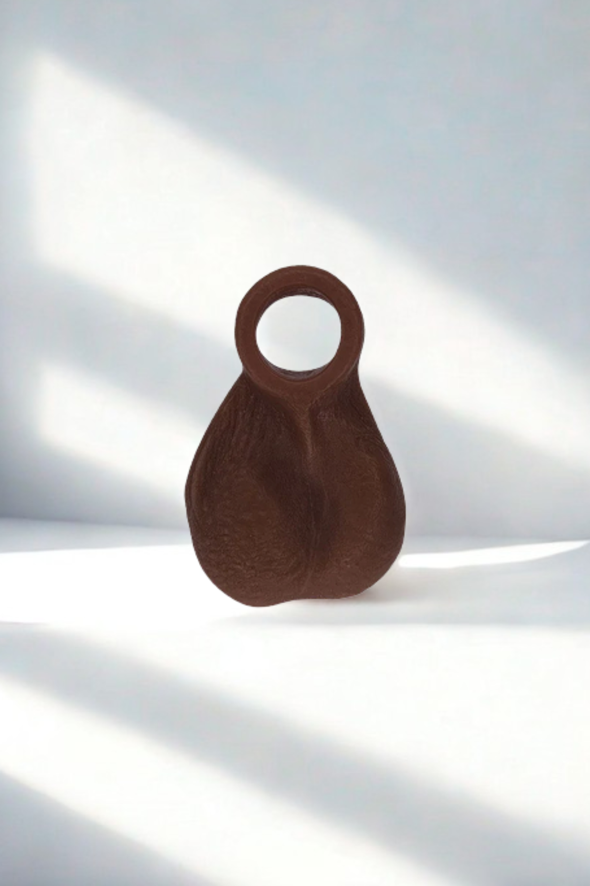 Ball Bump attachment cock ring in Chocolate standing against a white wall with soft sunlight and window shadows falling across it