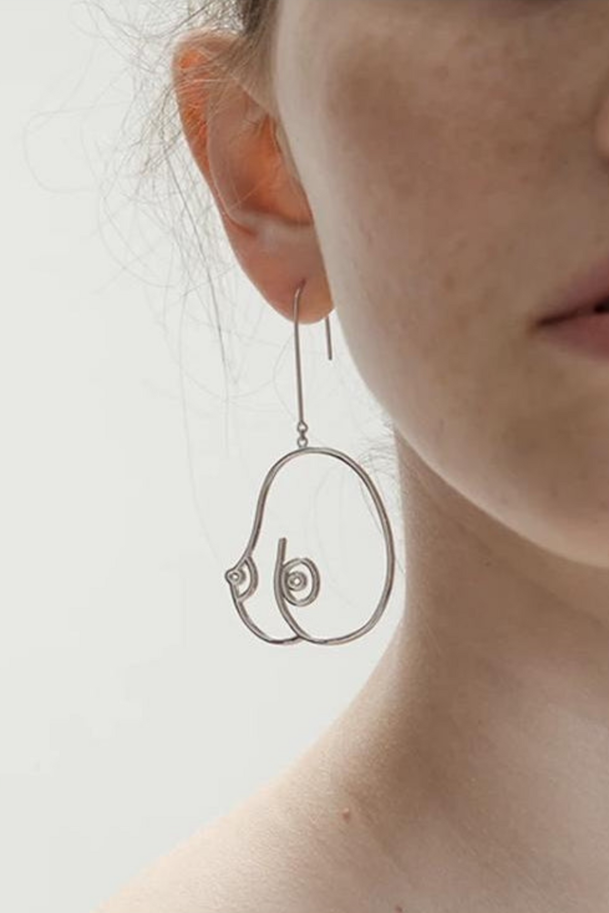 Close-up of a person wearing a silver wire earring shaped like a pair of breasts in outline, hanging from a long hook below the earlobe against a pale backdrop.