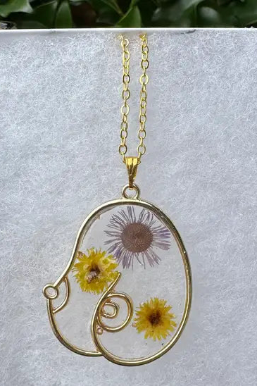 Boob Floral Resin Necklace in gold with clear resin holding pressed purple and yellow daisies, shown on the white cushion of its gift box