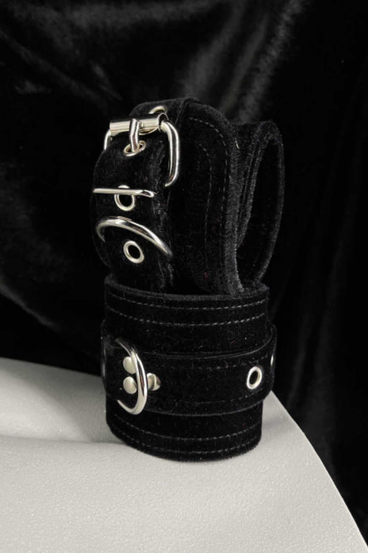 Black velvet D-ring cuffs with silver buckles stacked on a white mannequin form, set against a backdrop of draped black velvet fabric.