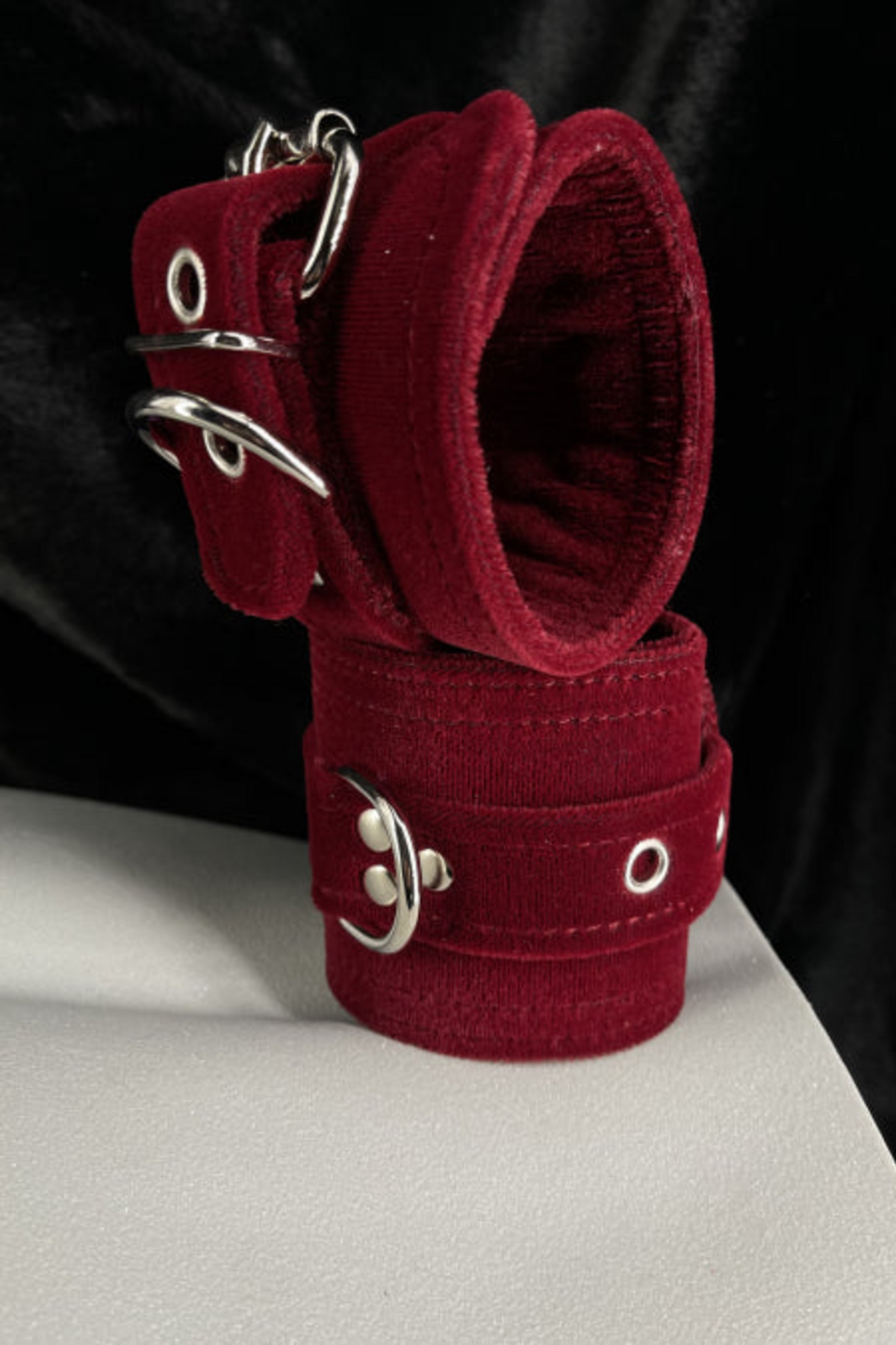 Burgundy velvet D-ring cuffs with silver buckles stacked on a white mannequin form, set against a backdrop of draped black velvet fabric.