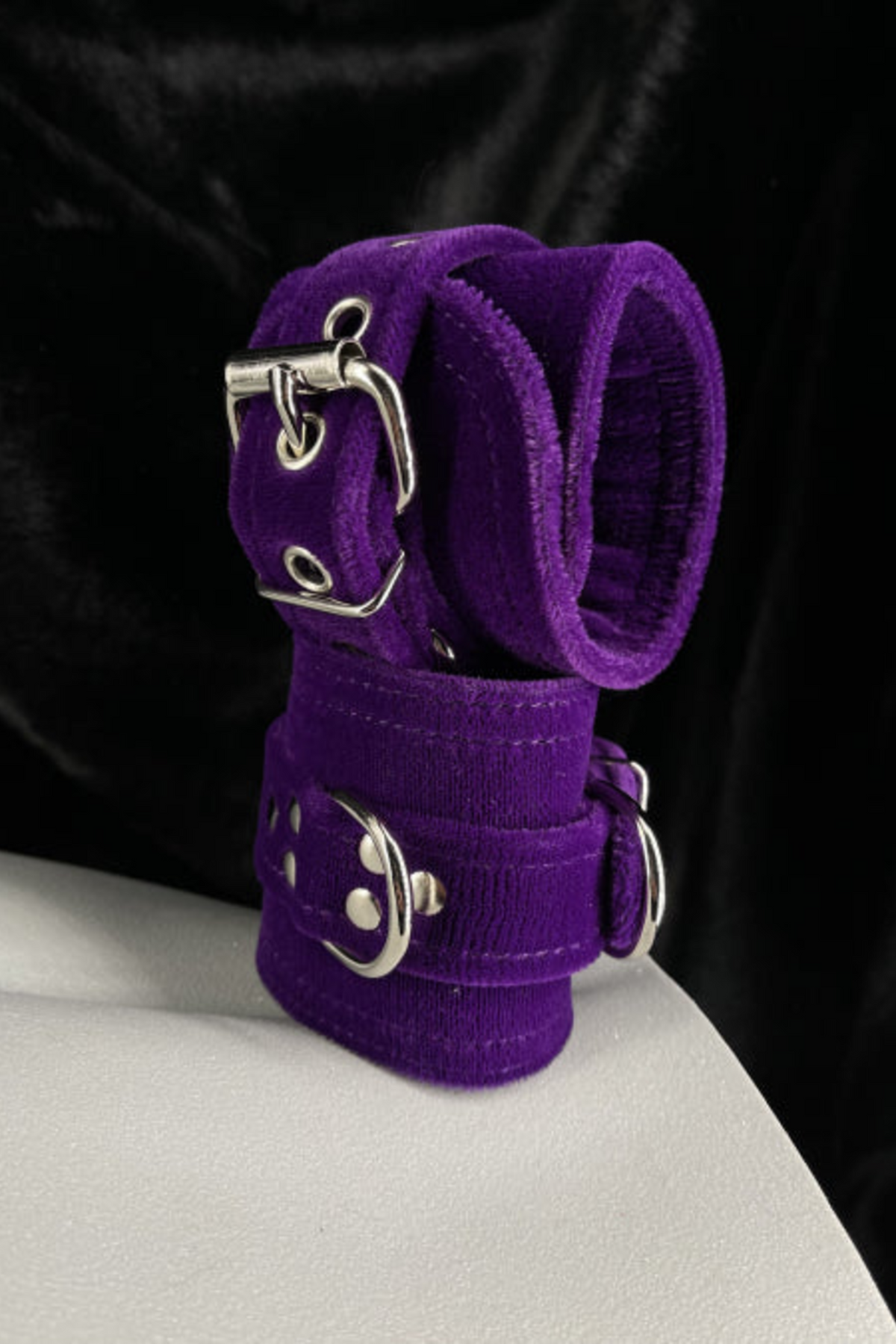 Purple velvet D-ring cuffs with silver buckles stacked on a white mannequin form, set against a backdrop of draped black velvet fabric.
