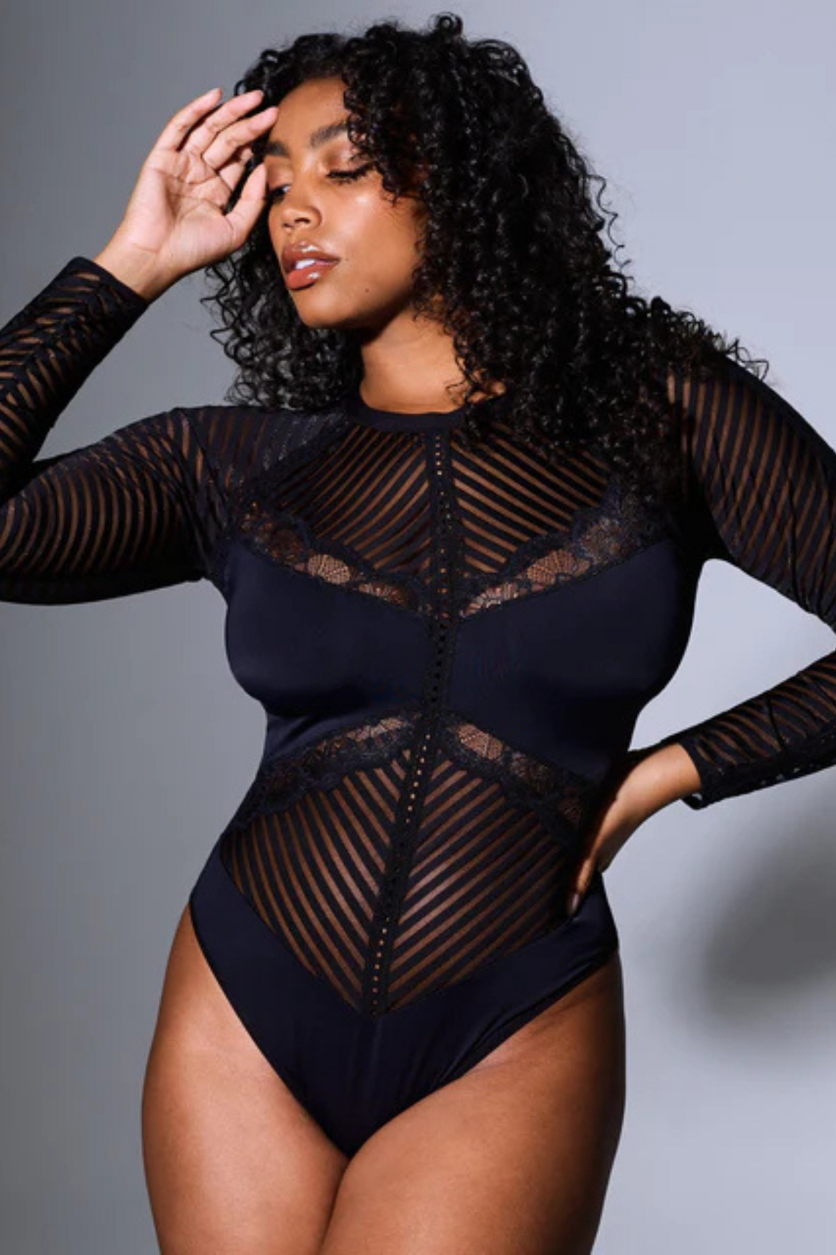 Plus-size model in Chelsea's High Line long-sleeved bodysuit in black with angled sheer stripe mesh panels and lace inserts