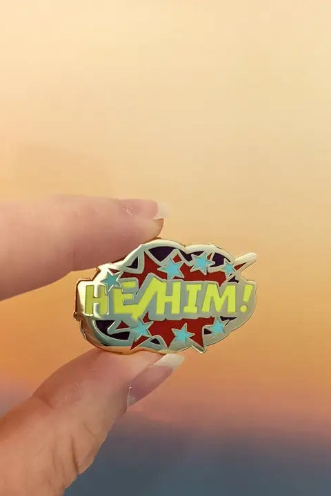 Fingers holding the Comic POW! He/Him enamel pin, a gold-edged comic burst with yellow-green lettering and stars, against a sunset sky