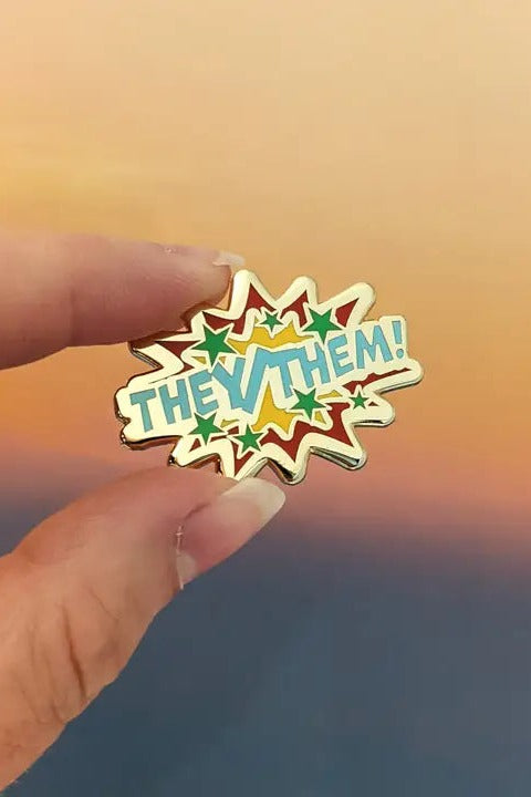 Fingers holding the Comic POW! They/Them enamel pin, a pale yellow starburst with teal lettering and colourful stars, against a sunset sky