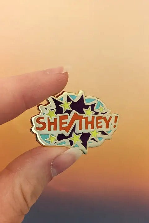 Fingers holding the Comic POW! She/They enamel pin, a pale blue burst with red lettering and dark purple stars, against a sunset sky
