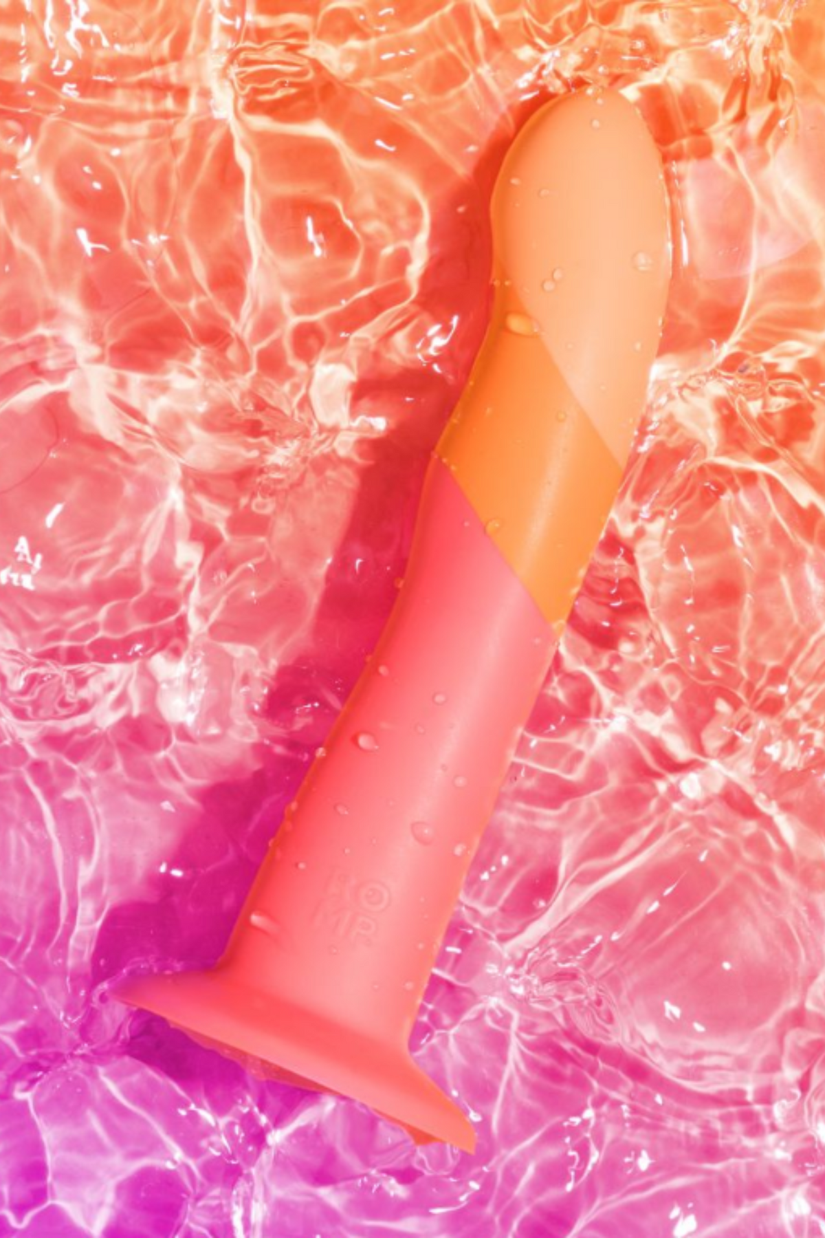 The coral and pink dildo standing in shallow rippling water, lit so the surface glows orange above and magenta below around its base.