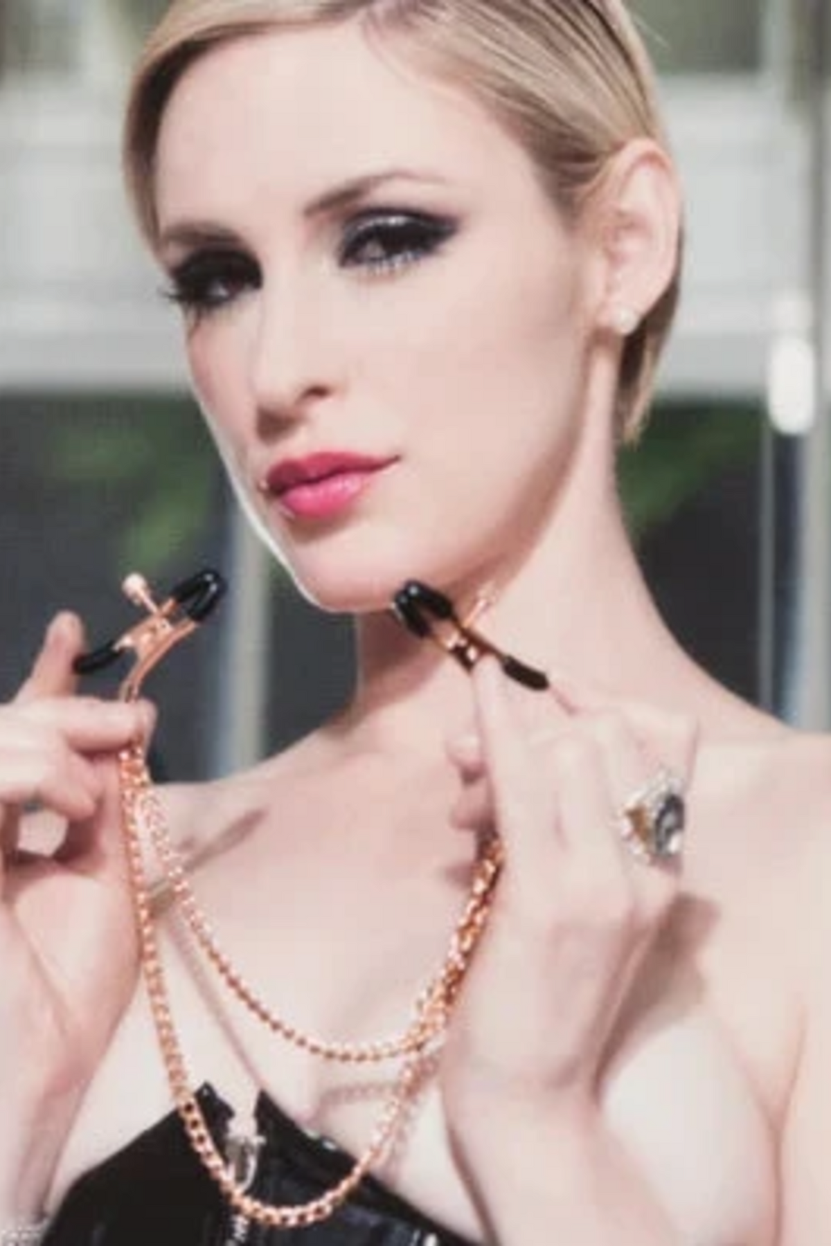 Model in a black top holds the rose gold nipple clamps up to her chest, the twin chains hanging in a loop between the two clamps.