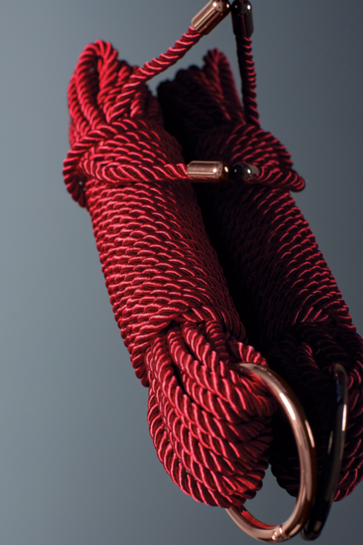 Coiled red silky bondage rope with a rose gold metal ring and rose gold end caps hanging against a dark grey gradient backdrop