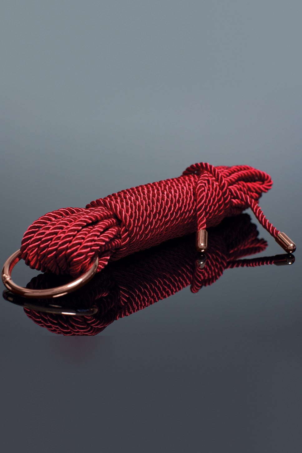 Hank of red twisted silky rope with a rose gold O-ring and two rose gold tips lying on a glossy dark surface with a mirror reflection below