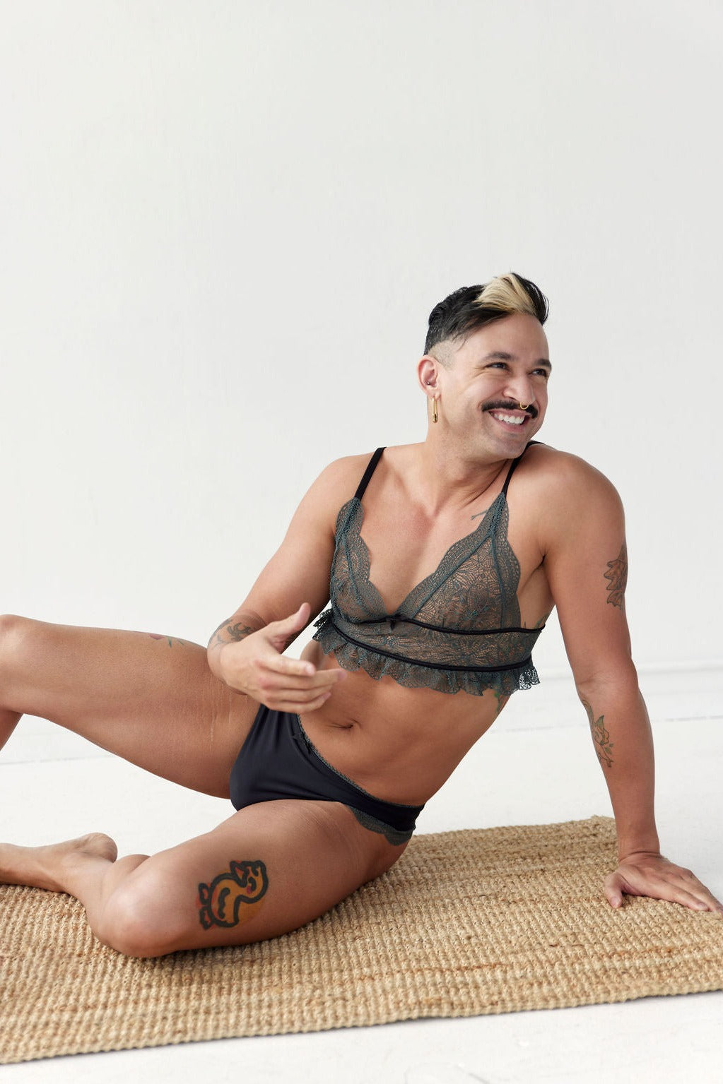 Smiling model seated on a jute rug in the Floruisse Lucy lace thong gaff in black with sage lace trim and a matching lace bralette