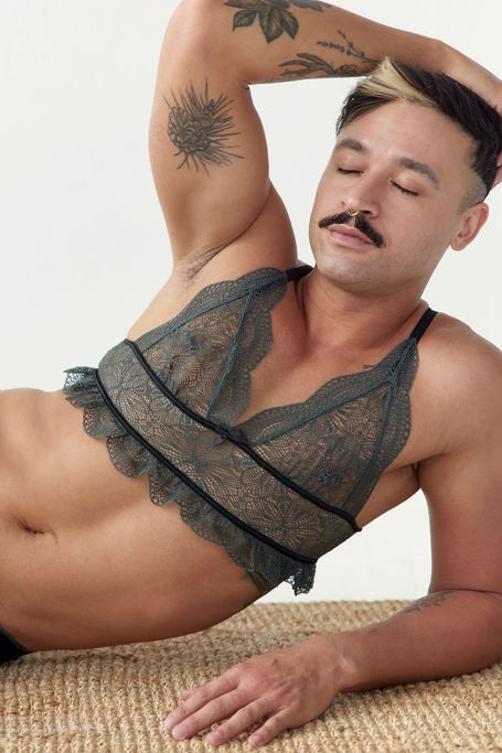 Reclining model with one arm raised wearing the Floruisse Ru lace bralette in slate sage, close crop on the gathered lace cups