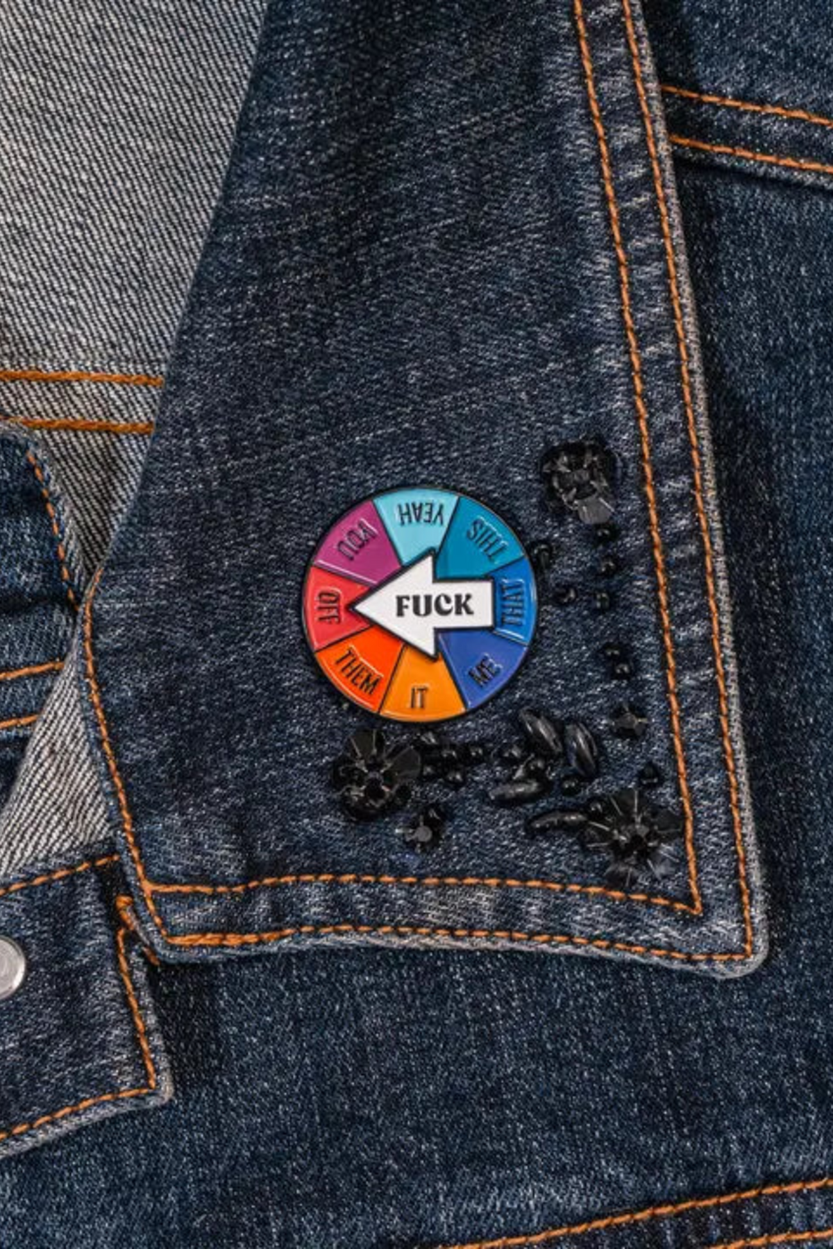 Round enamel spinner pin on a denim jacket collar, with a white arrow reading FUCK over a rainbow wheel of segments labeled you, me, them, off, it, yeah, this and that.