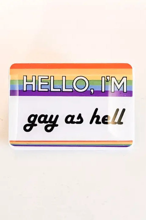 Rectangular enamel pin styled like a name tag with a rainbow stripe header reading Hello I'm and gay as hell in black script on a white body
