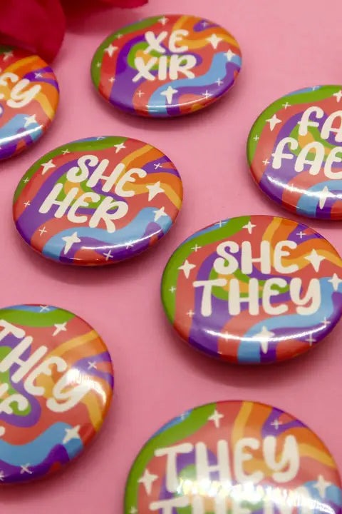 Scatter of rainbow holographic pronoun buttons on a pink surface, including She Her, She They, They She, Xe Xir and Fae Faer designs