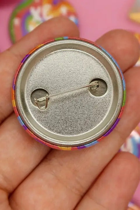 Back of a holographic rainbow pronoun button held in a palm, showing the silver metal backing and the straight pinback clasp