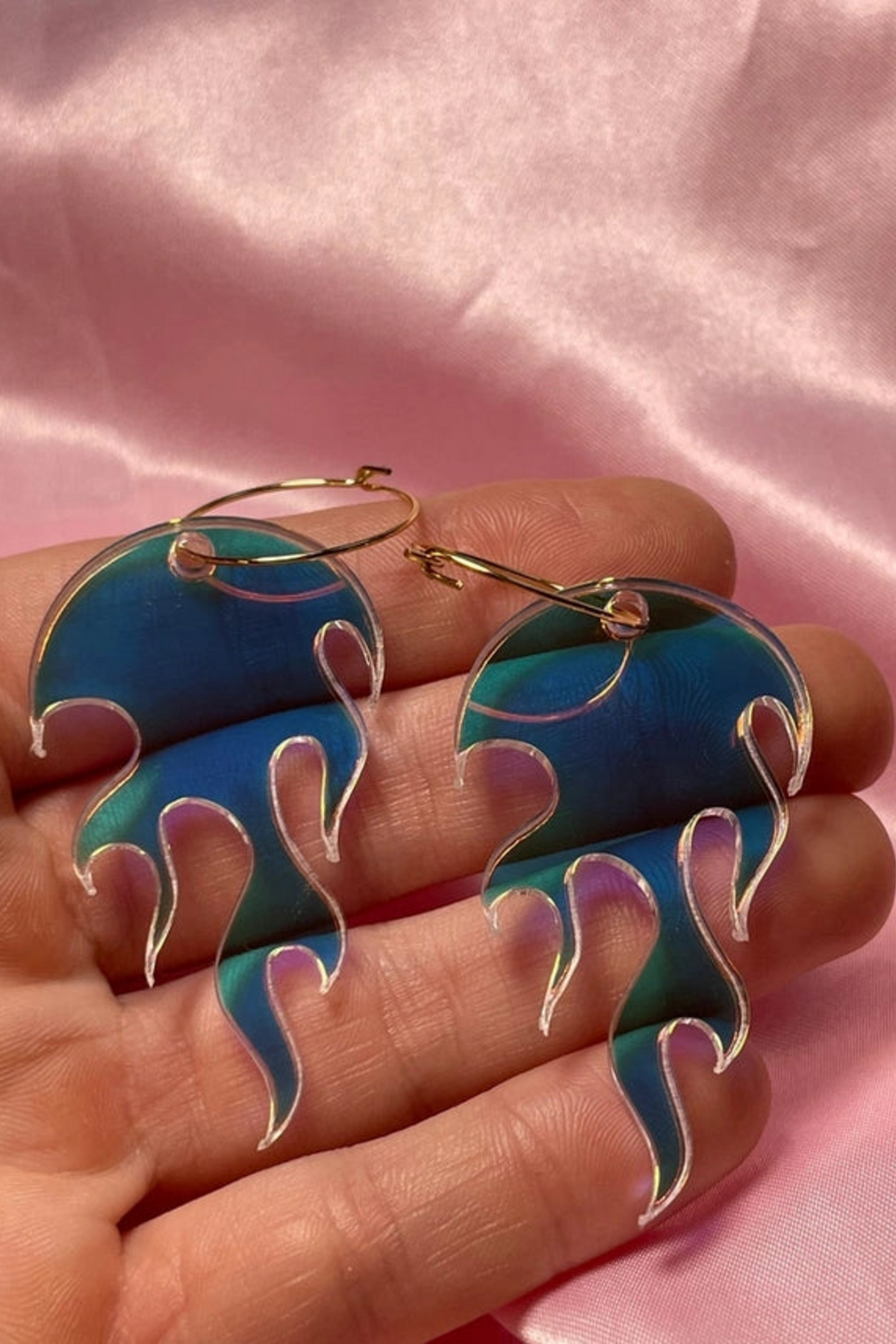Two iridescent flame shaped hoop earrings in blue and green resting on an open palm, photographed against a soft pink satin background.