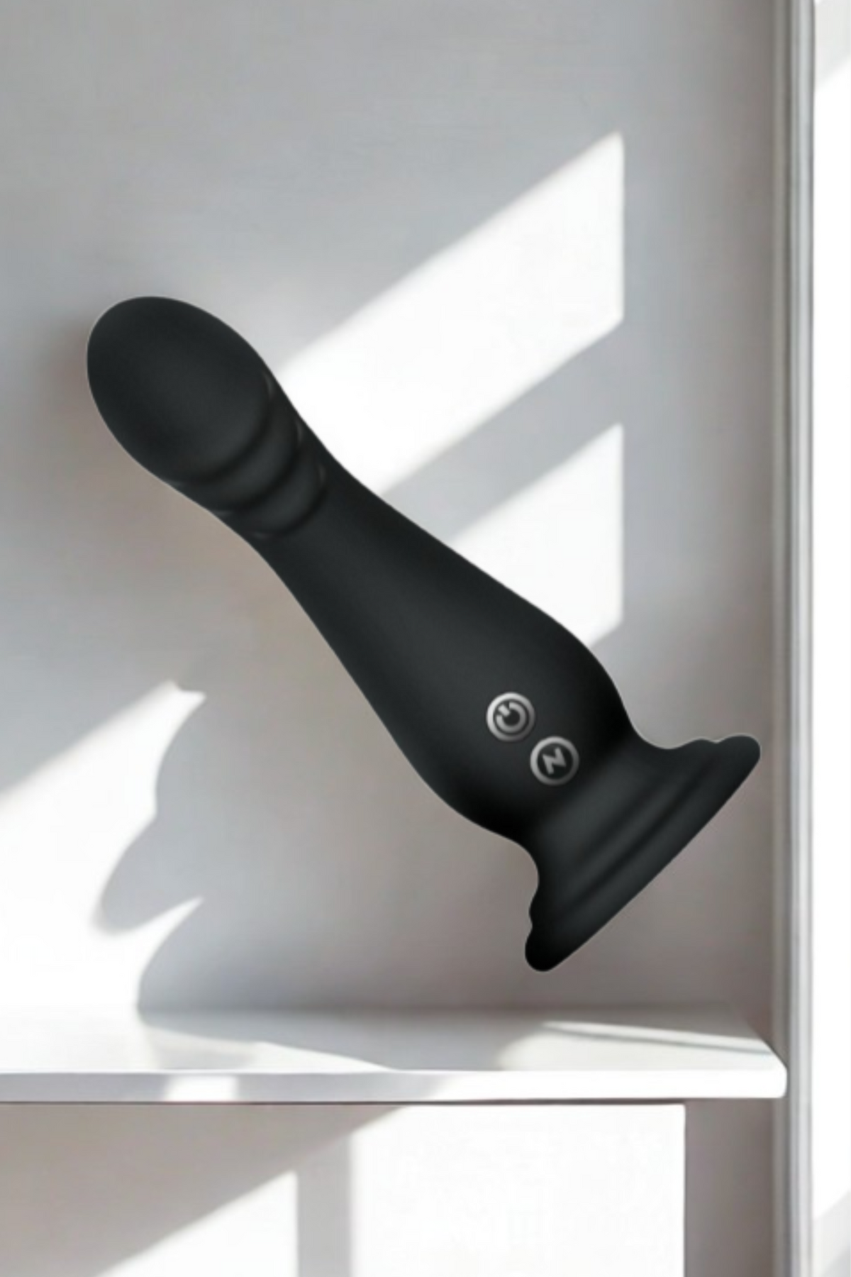 The black dildo resting on a white ledge, suction base to the right, with hard-edged window shadows falling across the wall behind it.