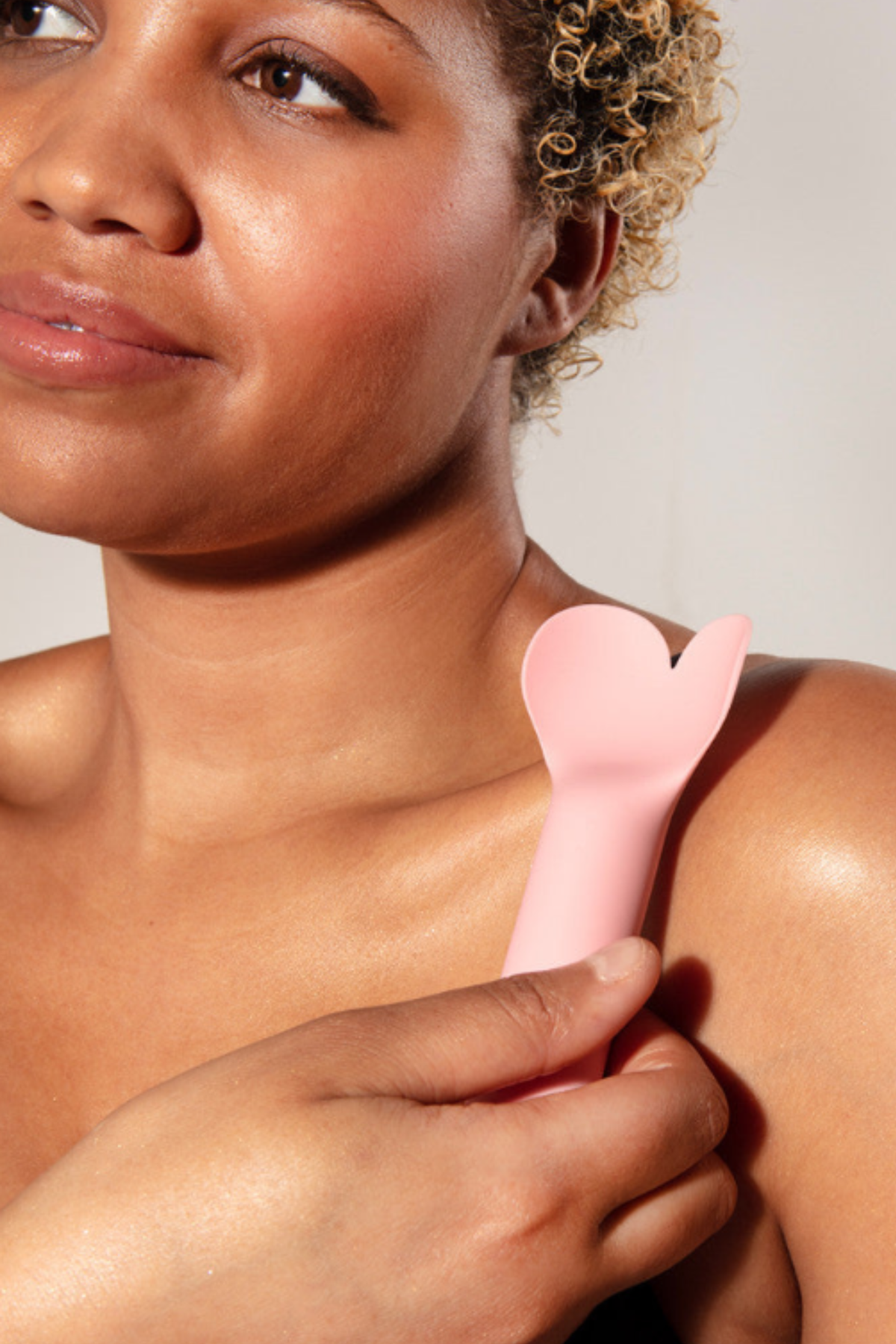 A smiling woman resting the dusty rose Amour bullet against her bare shoulder, holding it lightly by the handle with one hand.