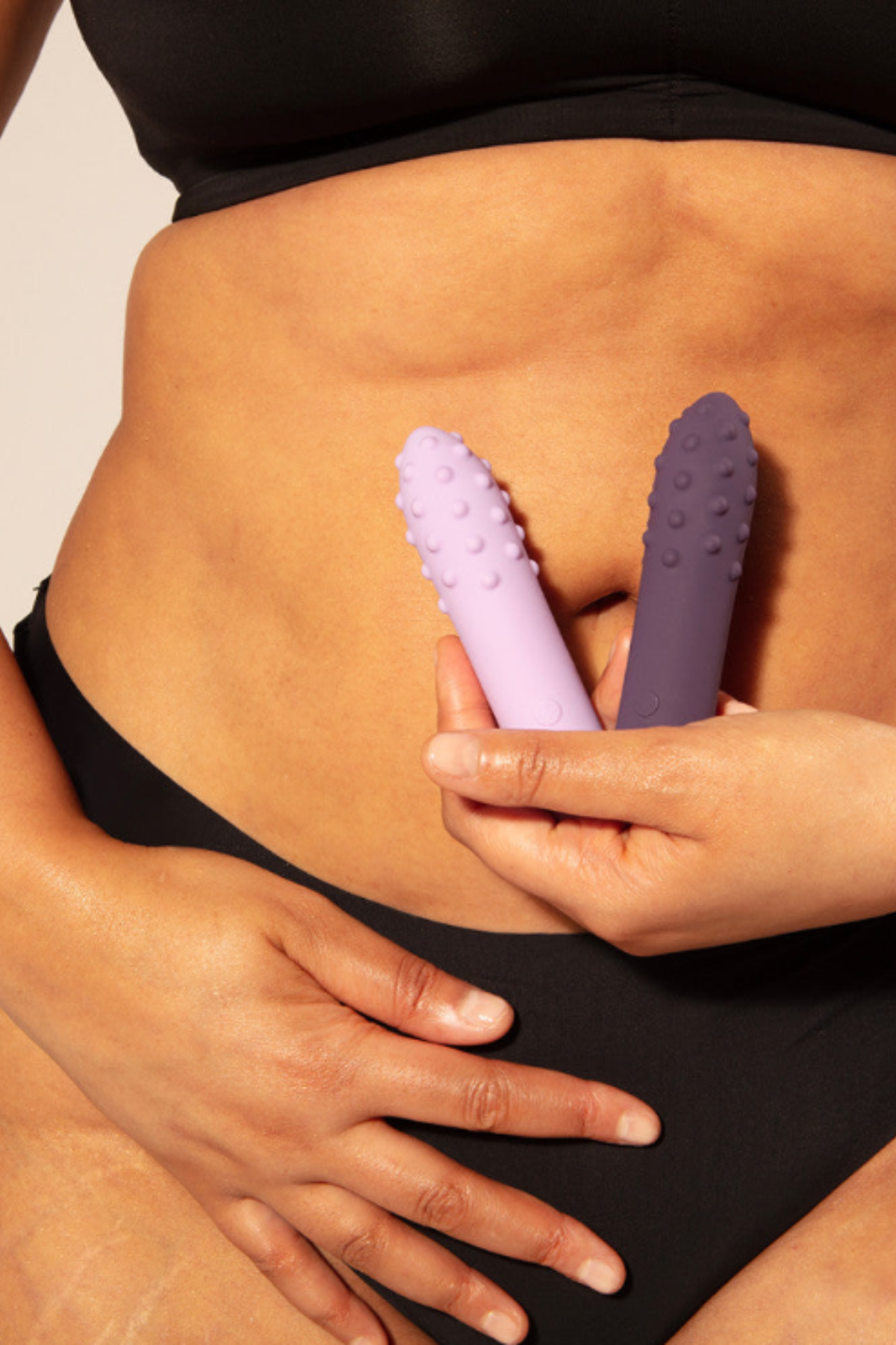 Model in black underwear holding the Je Joue Duet textured bullet vibrators in Lilac and Purple side by side against her lower stomach