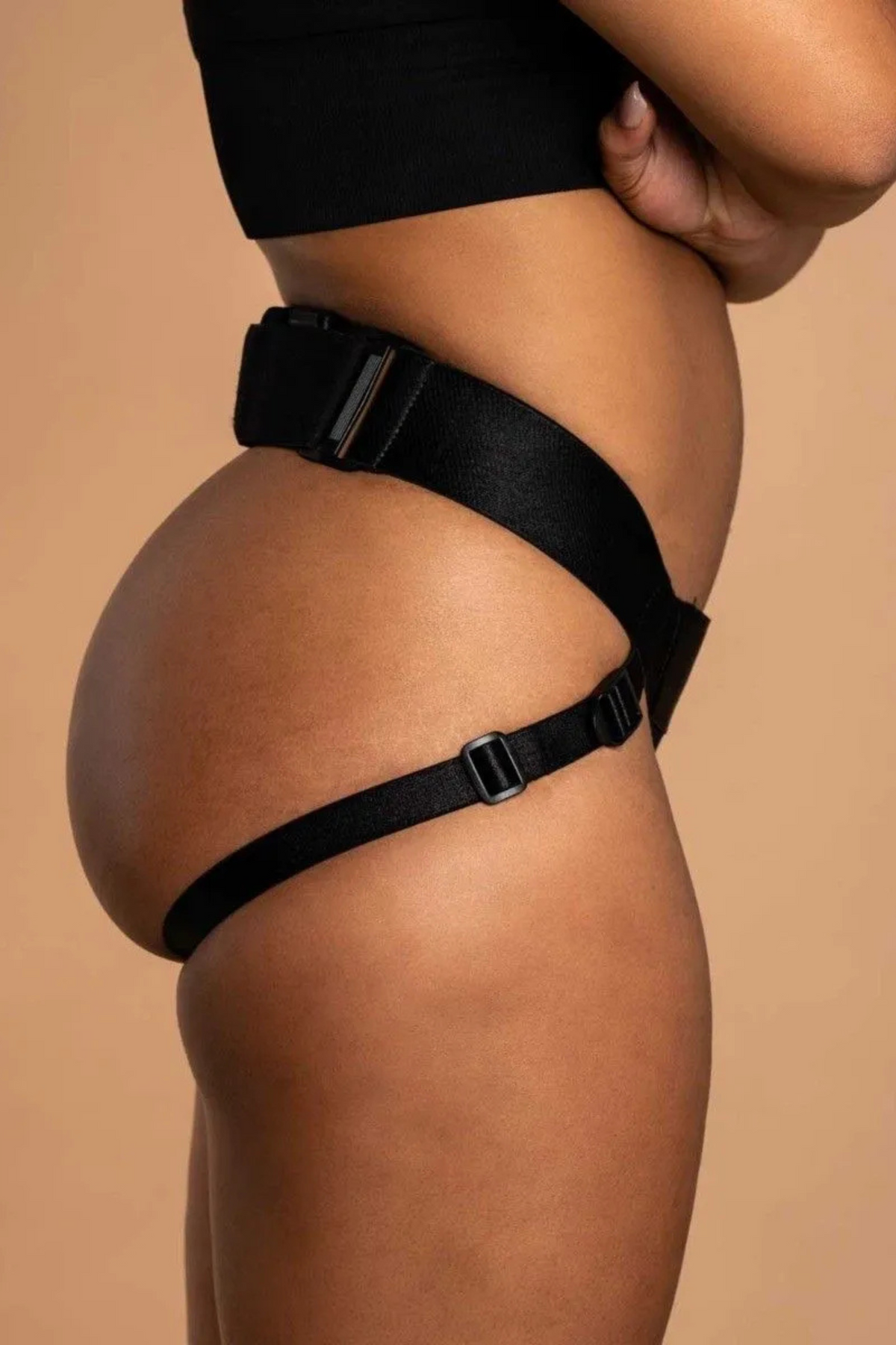Side view of a model wearing the black Jock open butt harness by Wet for Her, showing the waistband and thigh strap on a tan backdrop