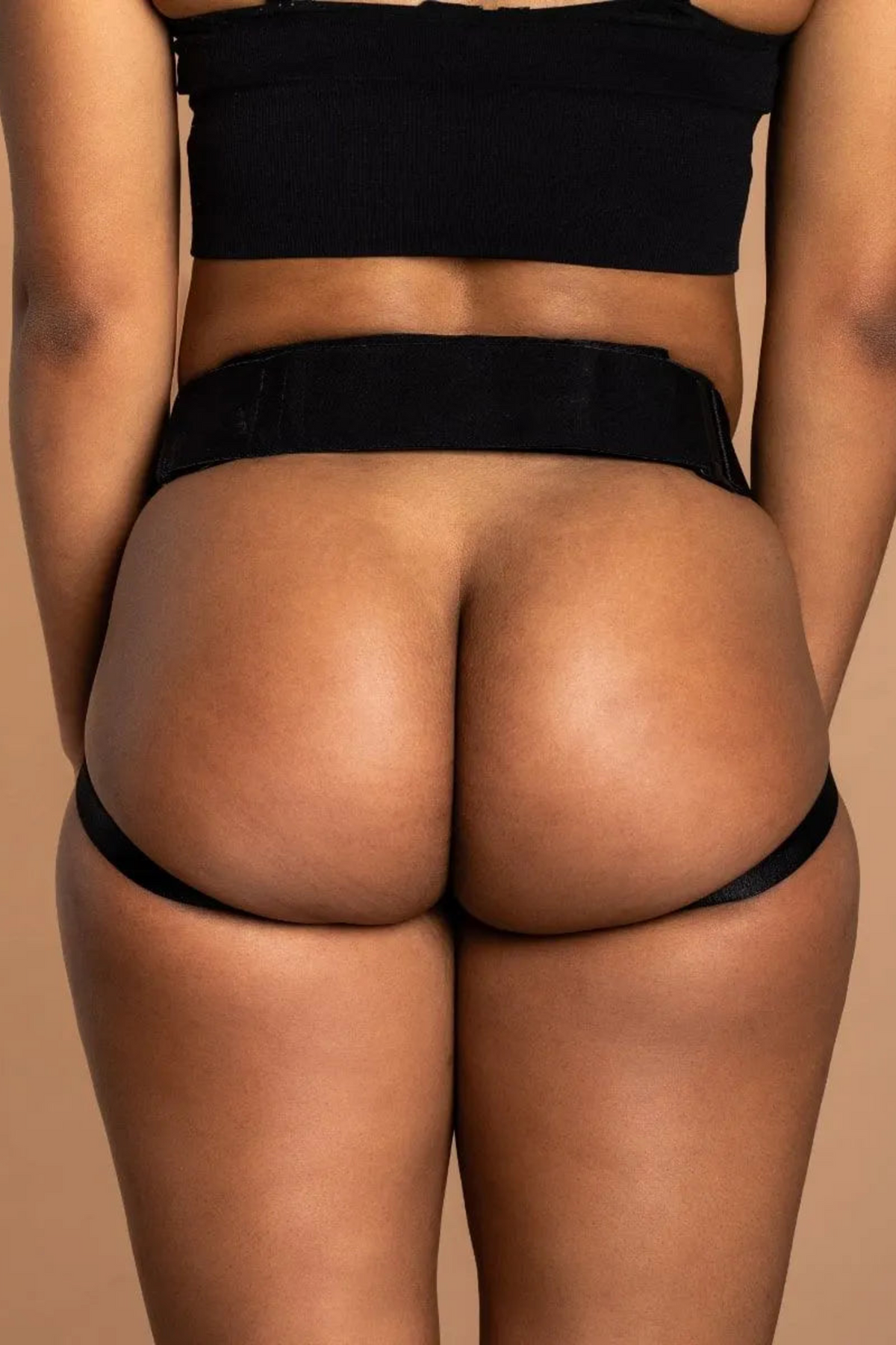 Back view of a model wearing the black Jock open butt harness by Wet for Her, showing the open back and thigh straps on a tan backdrop