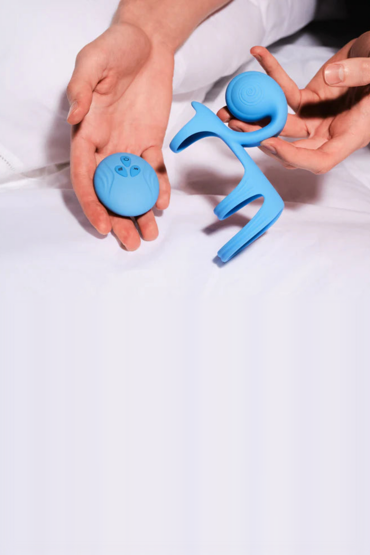 Hands on white sheets holding the blue Jovi Tri Arc Cock Ring and its round wireless remote, a lifestyle shot of the couples' toy in bed