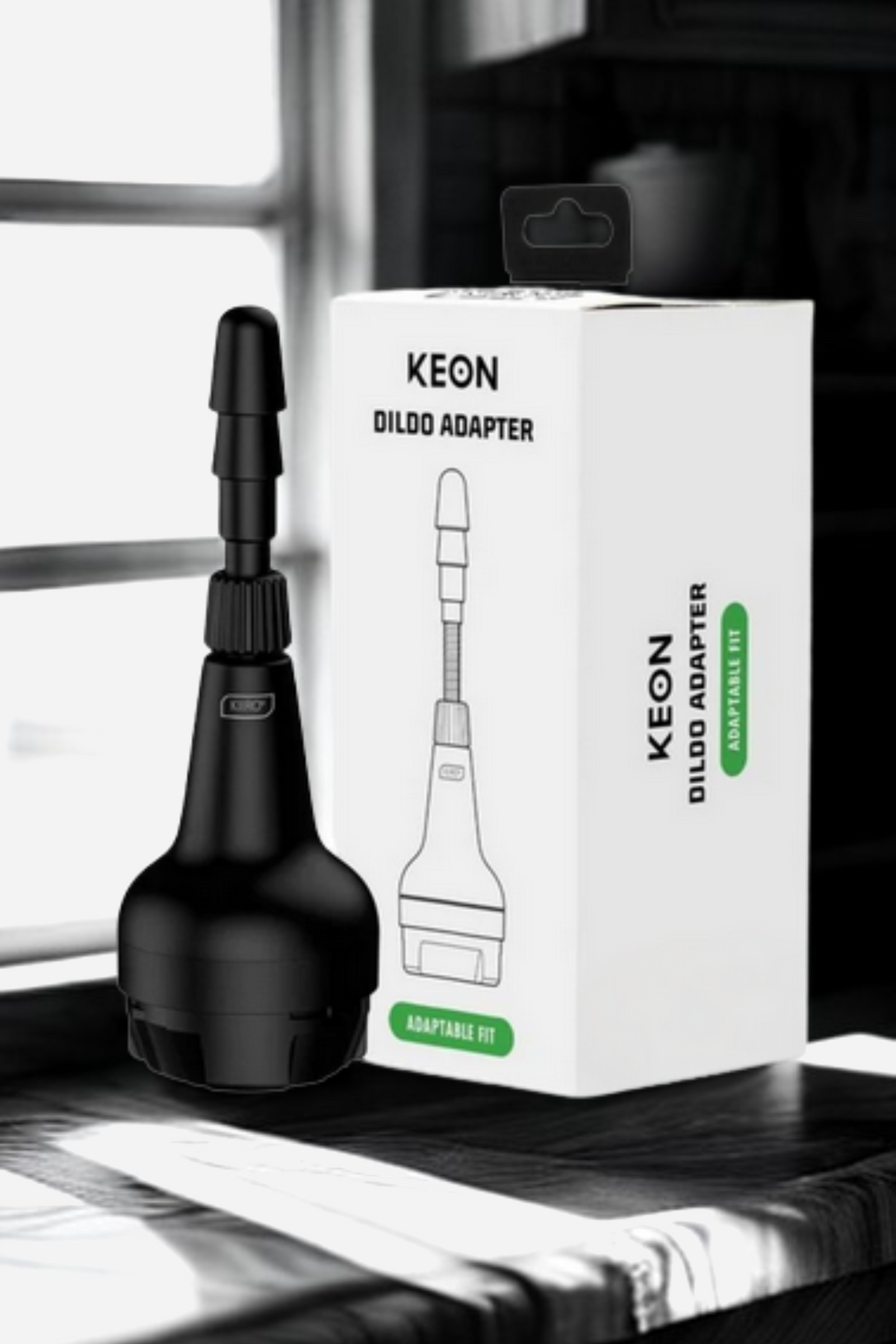 The black adapter standing beside its white retail box, which reads KEON Dildo Adapter, on a sunlit shelf with a bright window behind them.