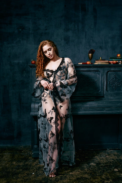 curly red haired model stands leaning next to a piano & looking down. She is wearing a regal mesh robe with black embroidery butterflies, the Kilo Brava Embroidery Maxi Robe in Black Butterflies