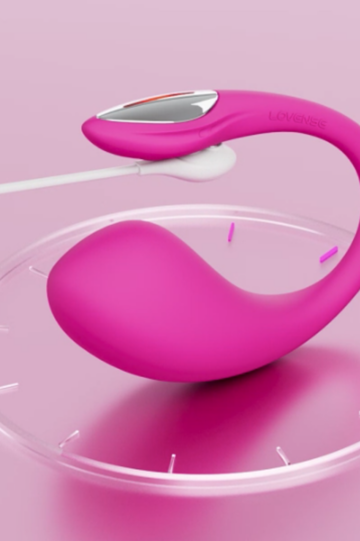 Hot pink Lovense Lush 4 resting on a clear glass stand against a soft pink studio backdrop, its egg-shaped bulb and curved antenna in close focus.