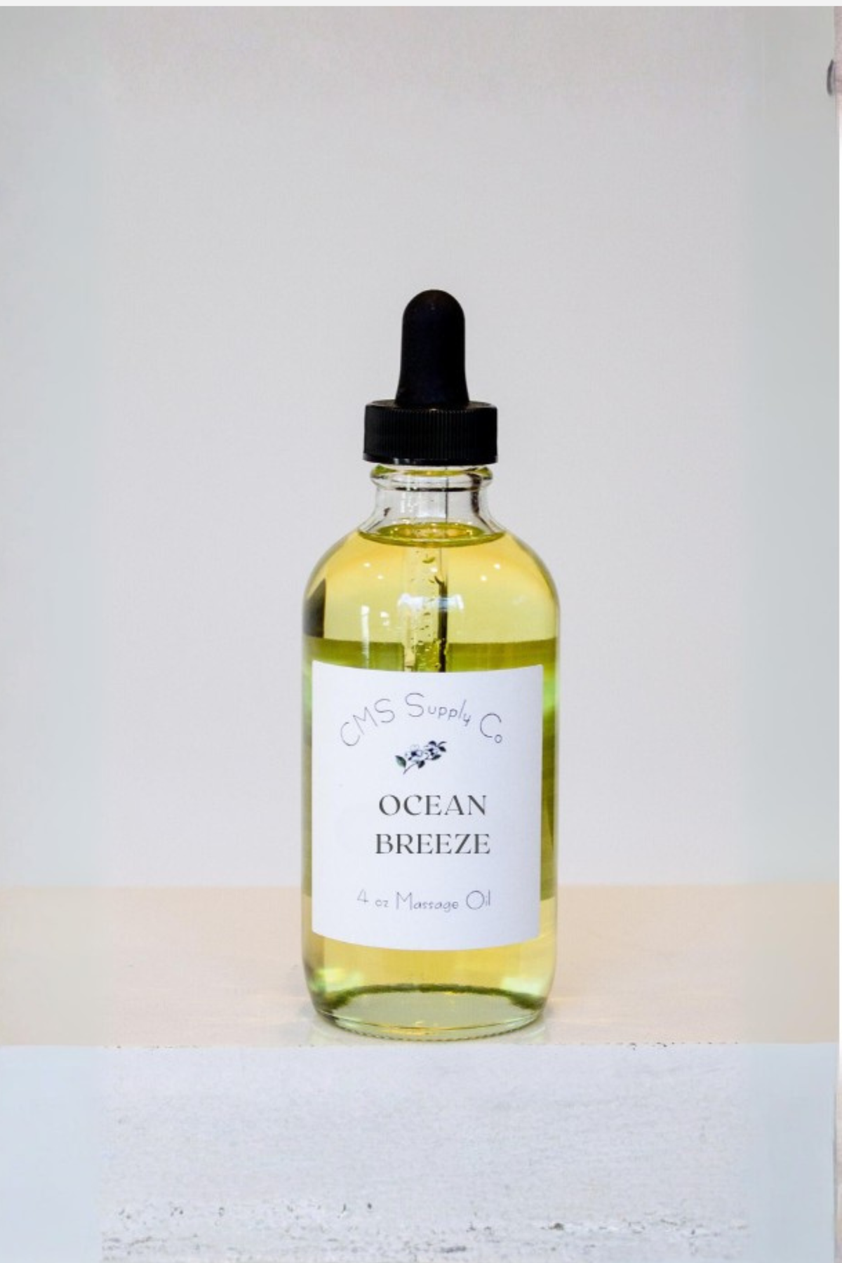 A 4 oz glass dropper bottle of CMS Supply Co Ocean Breeze massage oil with a white label, standing on a white table