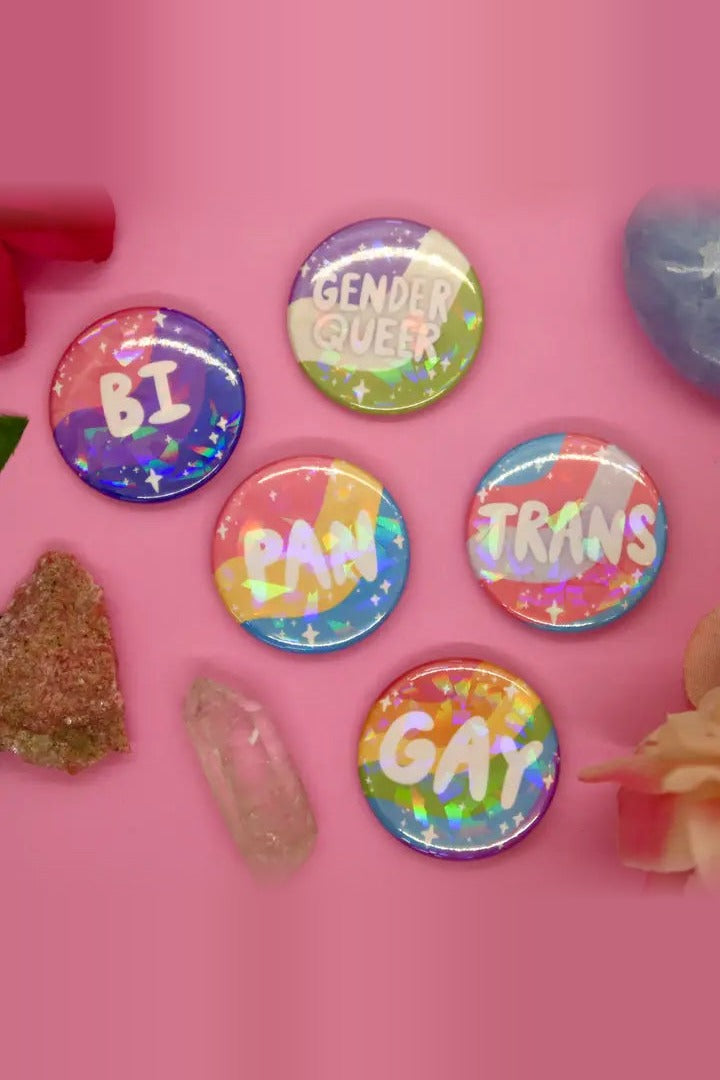 Bi, Genderqueer, Pan, Trans and Gay identity buttons shown in a holographic finish on pink; the buttons sold are not holographic
