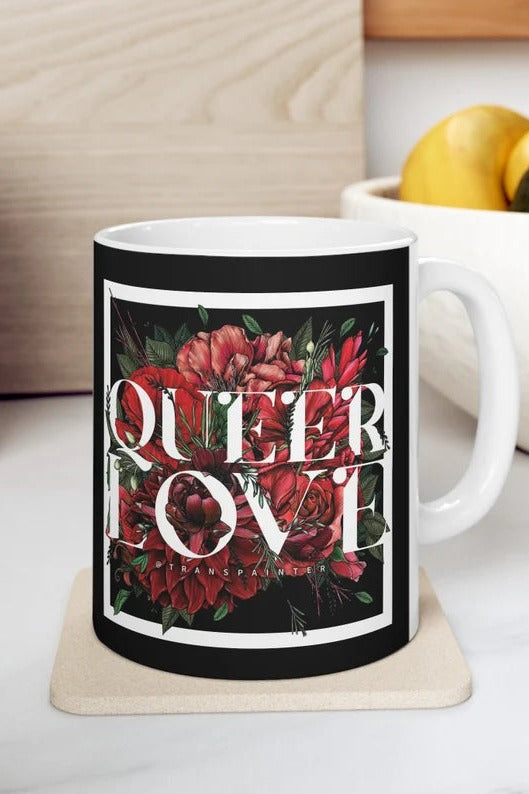 White ceramic mug with a black printed wrap reading Queer Love in white serif letters over red roses, on a wooden coaster beside a bowl of lemons