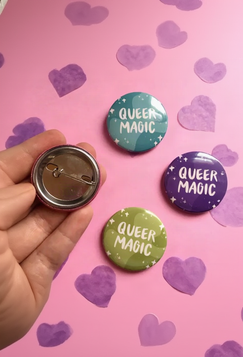 Queer Magic buttons in teal, purple and green on pink with paper hearts, and a hand holding one to show the metal pin back