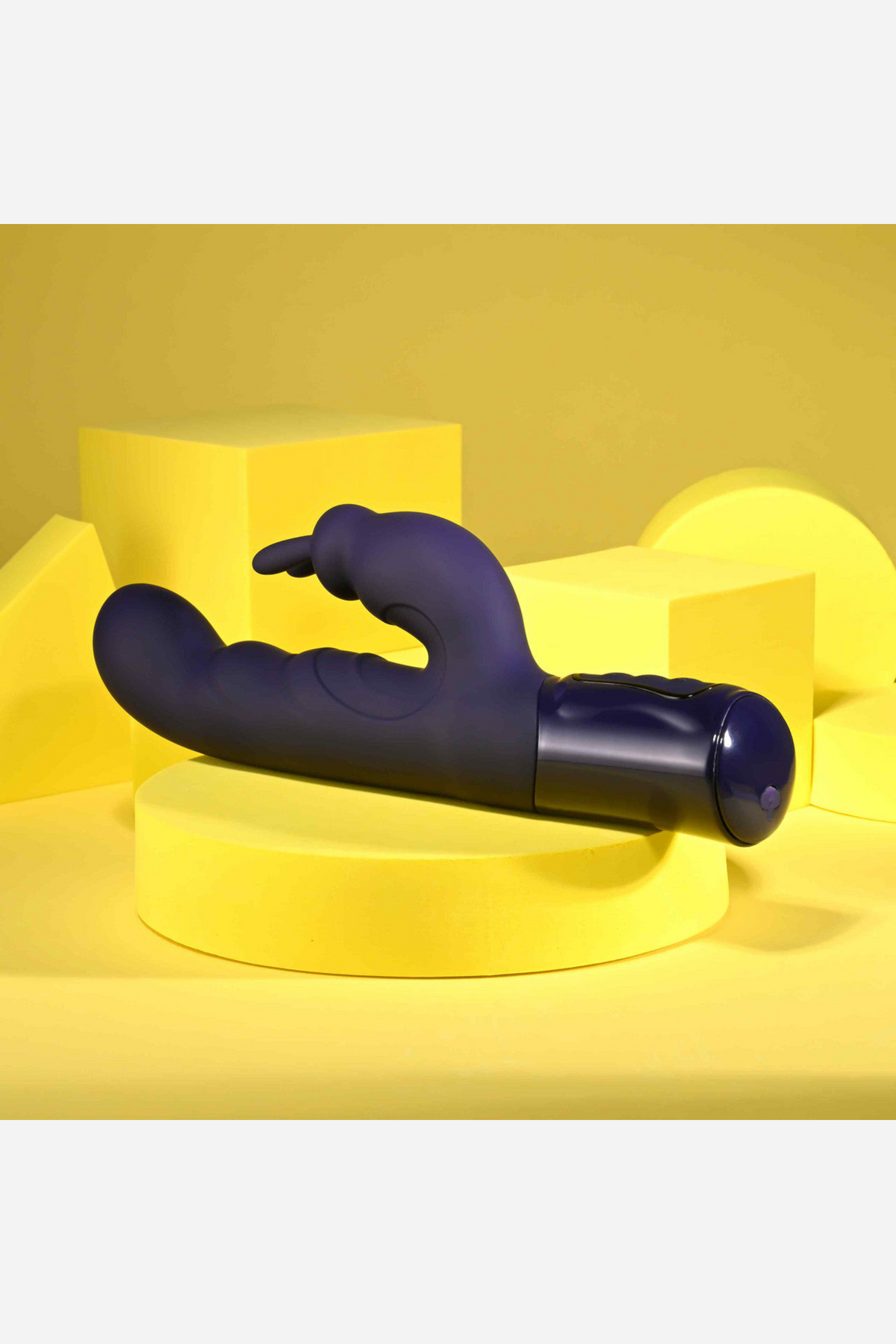 Purple She's A Big Deal Rabbit Vibe resting on yellow studio blocks, a side view of the curved dual motor shaft and flexible clitoral arm, on grey