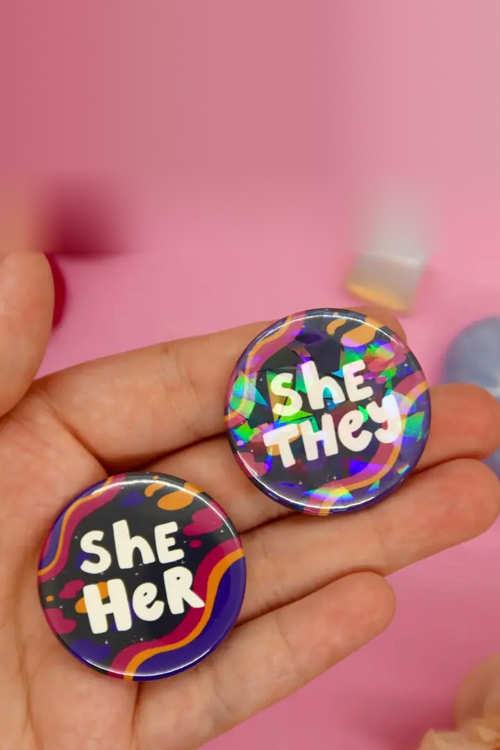 Hand holding two holographic Spacey pronoun buttons reading She/They and She/Her in white bubble letters over wavy stripes on pink
