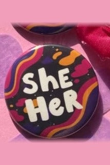 Spacey She/Her pronoun button with white bubble lettering over orange, pink and purple wavy stripes on a dark background, on pink felt