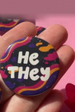 Hand holding the Spacey He/They pronoun button with white bubble lettering over wavy orange, pink and purple stripes on a dark background