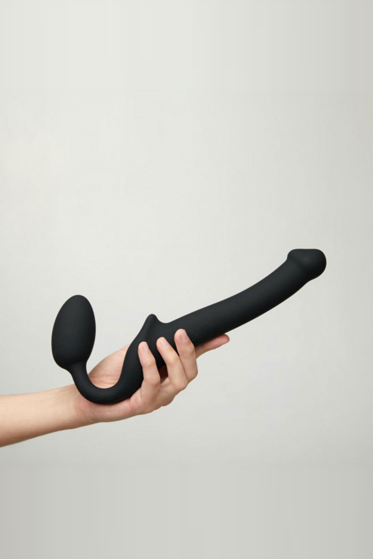 Strap-On-Me adjustable strapless dildo in black held in one hand against a soft grey wall, showing its long bendable shaft and bulb