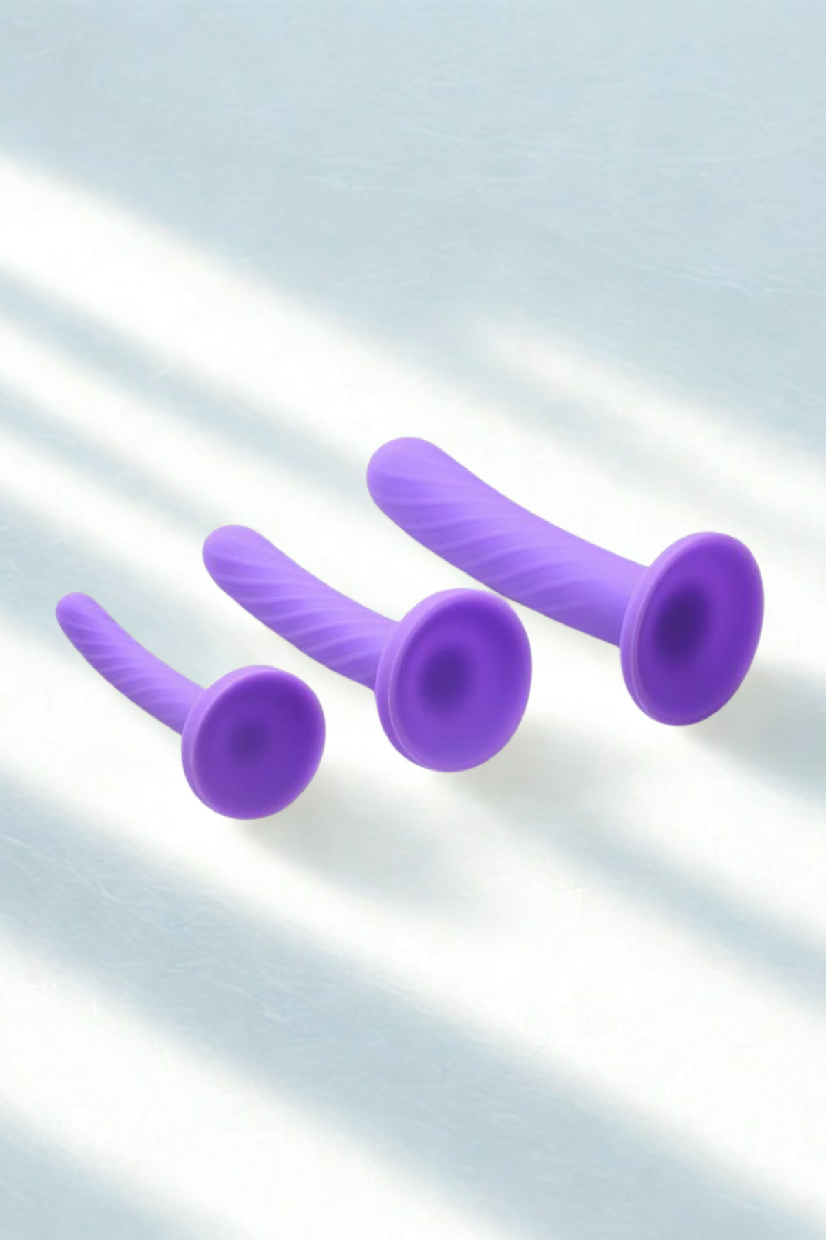 The three purple dildos lying on a pale surface with soft diagonal window shadows falling across it, their flared bases turned towards the camera.