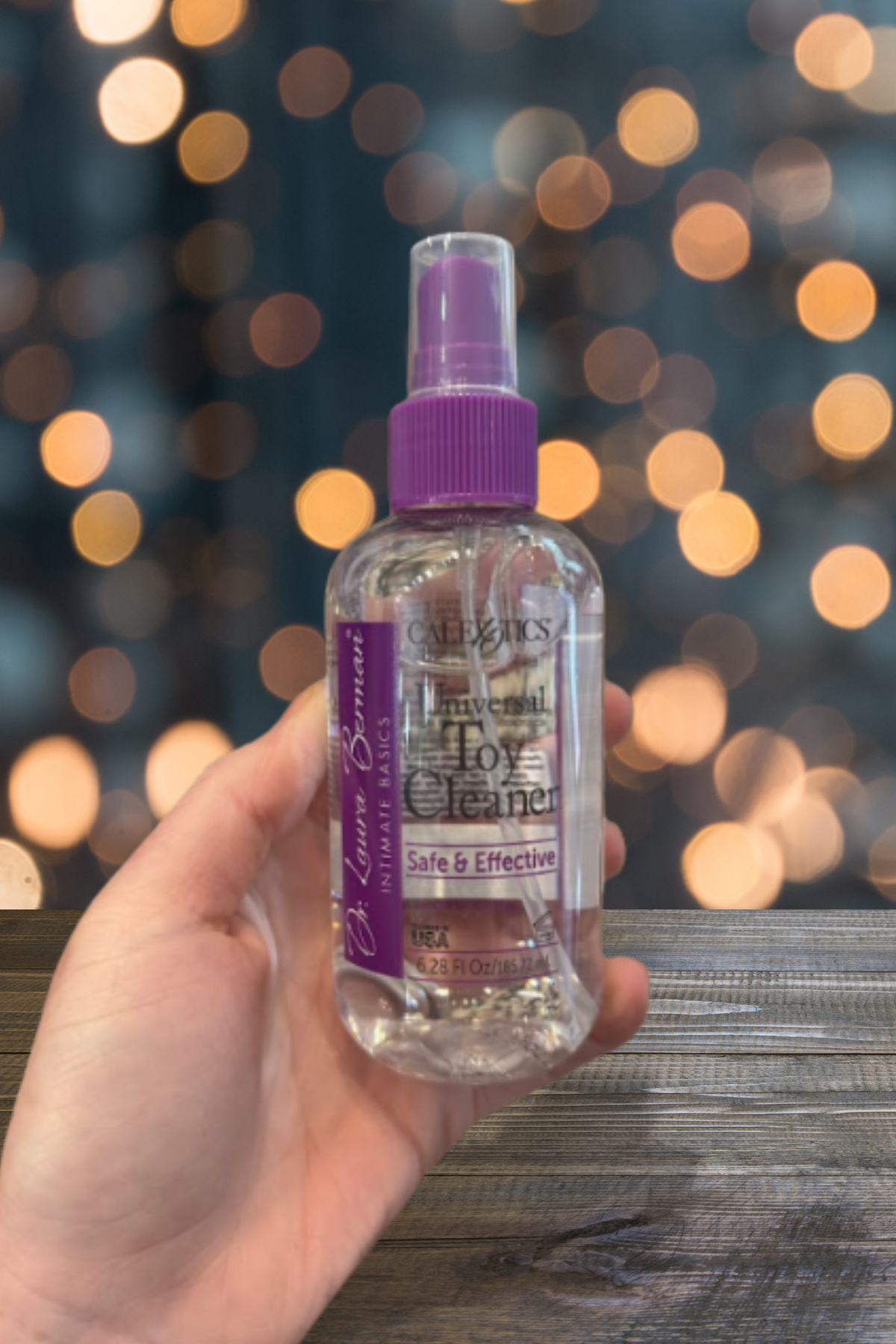A hand holds the toy cleaner bottle above a weathered wooden rail at dusk, with warm orange bokeh lights blurred in the background behind it.