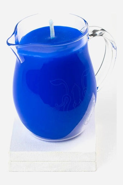 Blue wax play pitcher candle in a clear glass jug with handle and pour spout, set on a small white block against light grey