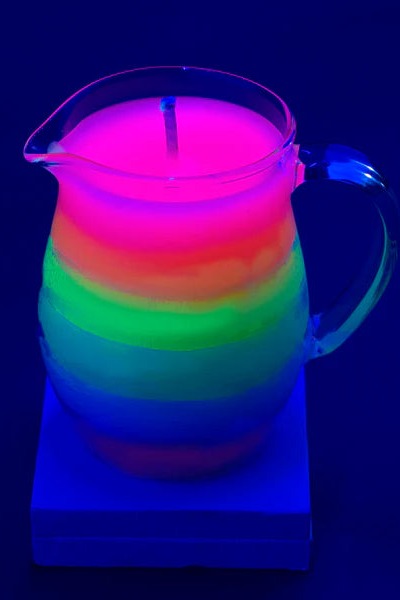 Rainbow wax play pitcher candle glowing neon pink, green and blue under UV black light, set on a small block against a dark blue backdrop