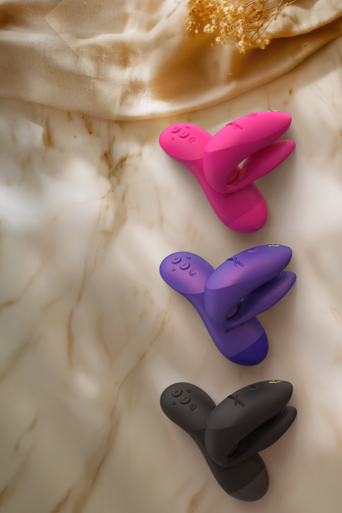 We-Vibe Chorus Pro couples vibrators in Hot Pink, Cosmic Purple and Black lying on draped cream fabric with gold accents