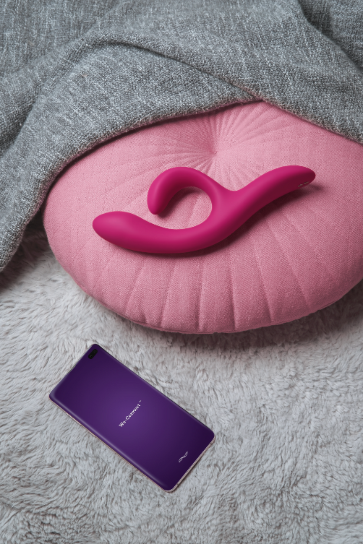 Magenta We-Vibe Nova 2 resting on a pink cushion over a soft grey throw, with a phone running the We-Vibe app to control it remotely