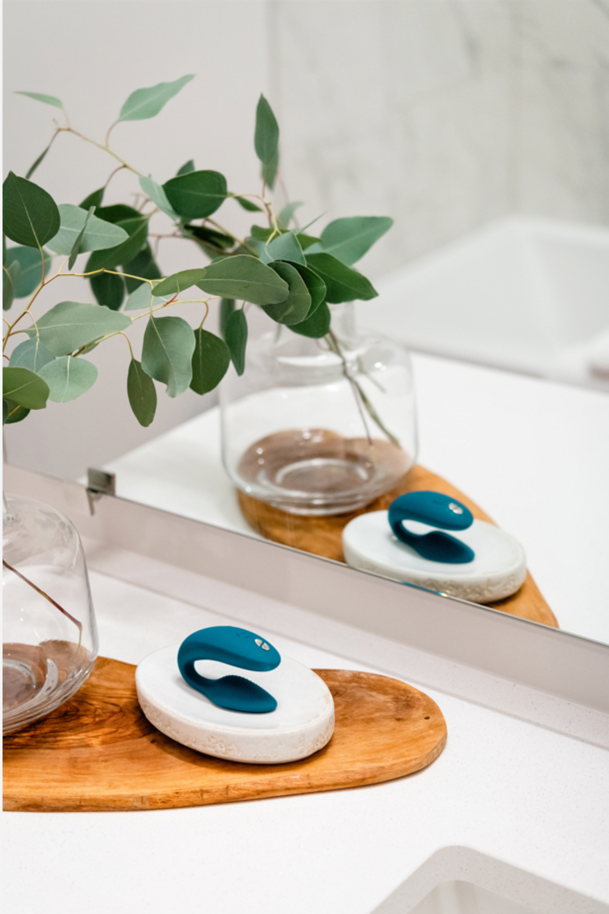Green velvet We-Vibe Sync 2 resting in its charging case on a wooden tray by a bathroom mirror, a lifestyle shot with fresh greenery